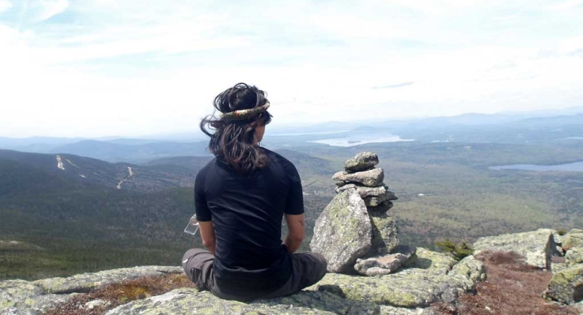 A person sits on an overlook at a high elevation and looks out at the vast, forested landscape below. 